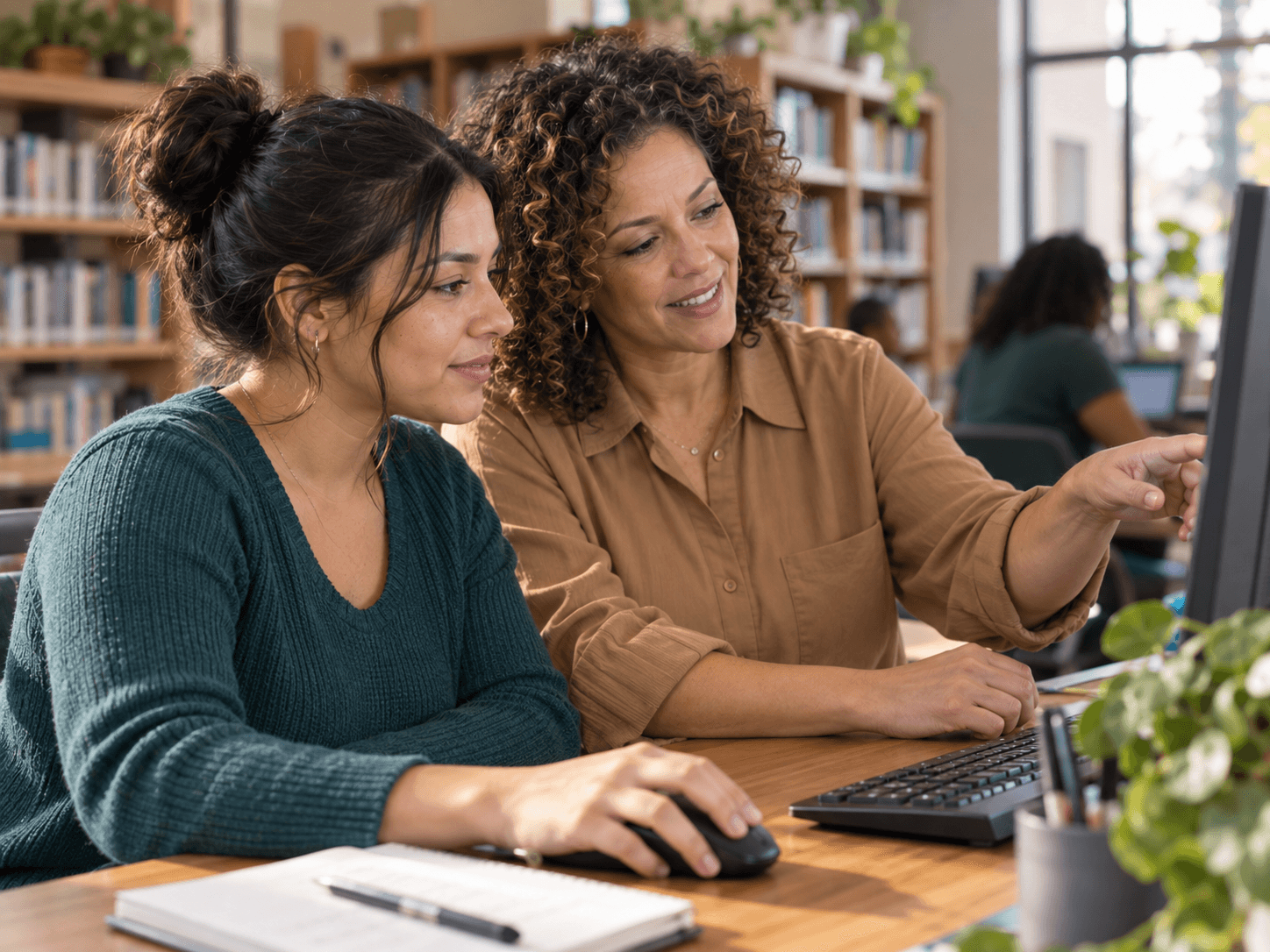 A woman helping a young adult learn a computer skill in a library